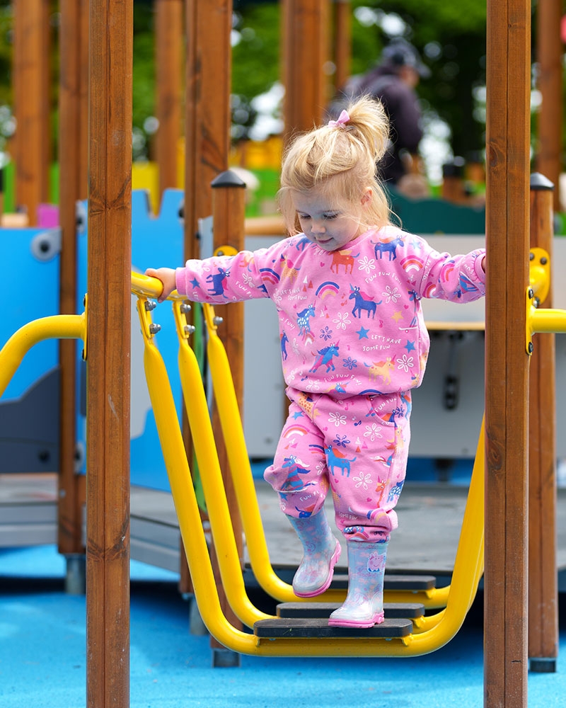 Ein Kleinkind in heller Kleidung überquert konzentriert eine Brücke auf einem Spielplatz.